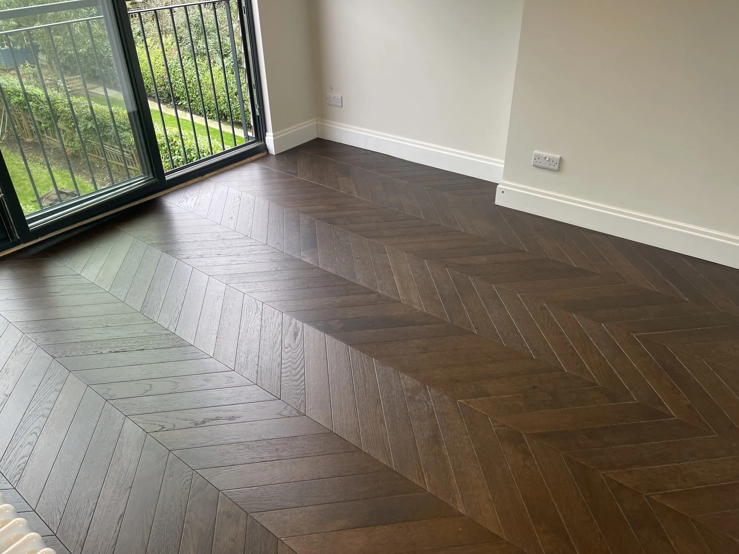 Dark engineered herringbone flooring in a bright room with garden views, installed by Woodford Flooring Ltd in Chigwell.