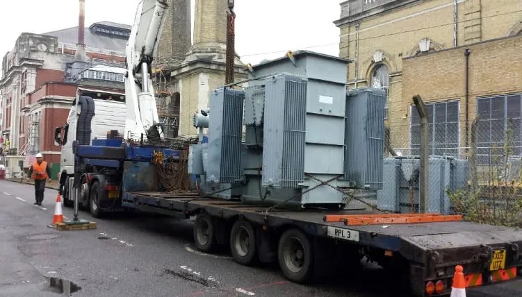 A large industrial transformer being loaded onto a flatbed truck, illustrating heavy equipment recycling by London Metal &amp; Recycling Ltd in Barking.