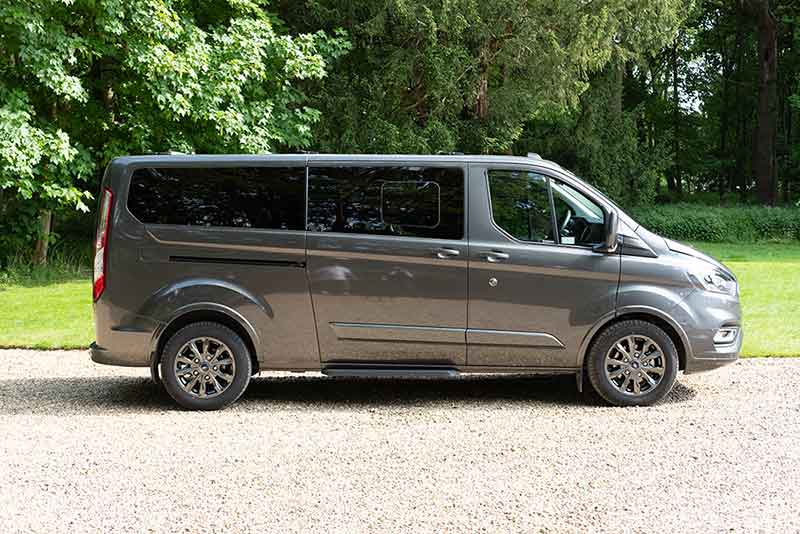 A grey minibus parked on a gravel path with trees in the background, available for 9 Or 17 Seater Minibus For Hire from Alley Cat Car &amp; Van Rentals in Whittlesford.
