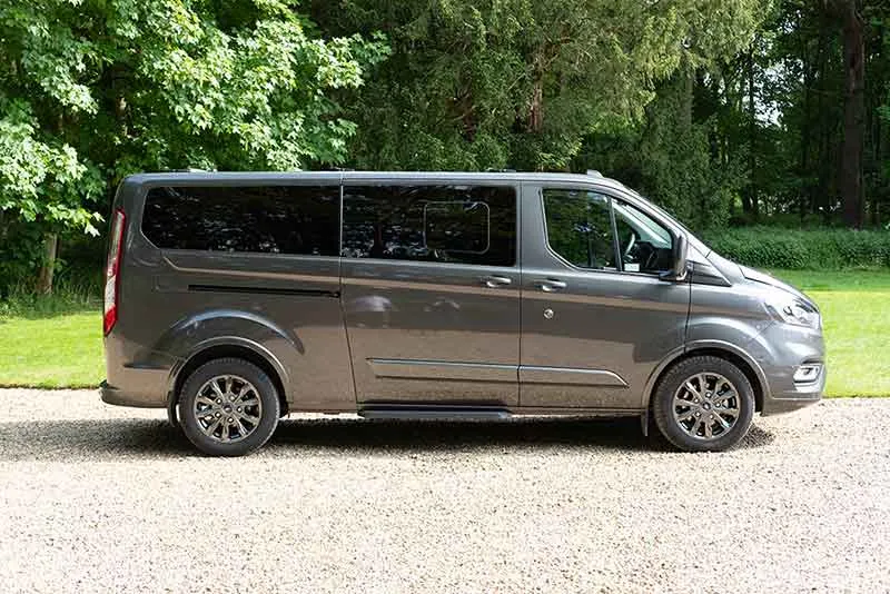 A grey minibus parked on a gravel path with trees in the background, available for 9 Or 17 Seater Minibus For Hire from Alley Cat Car &amp; Van Rentals in Sawston.